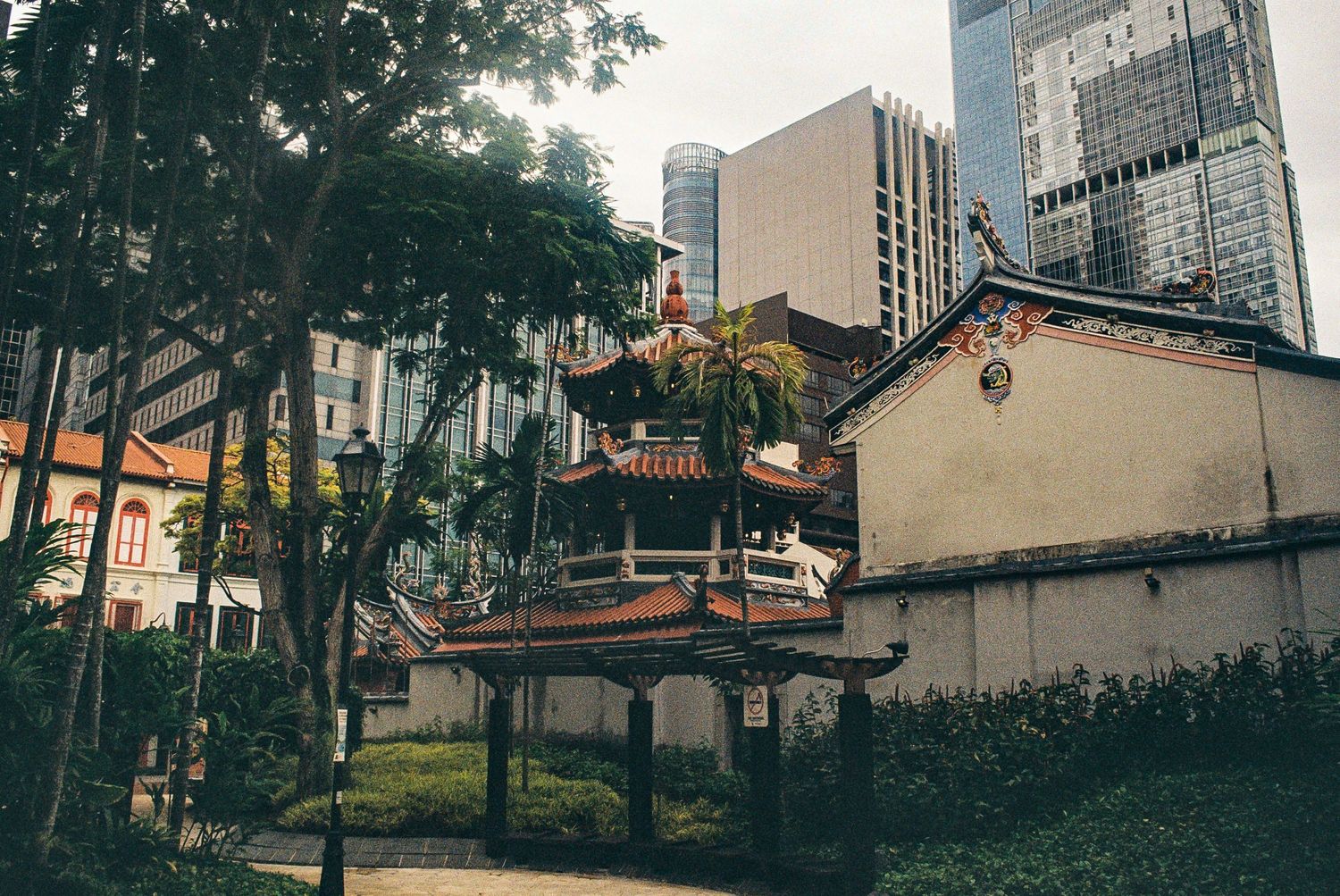 Old traditional architecture contrasts with modern skyscrapers in an urban park setting with lush trees and greenery.