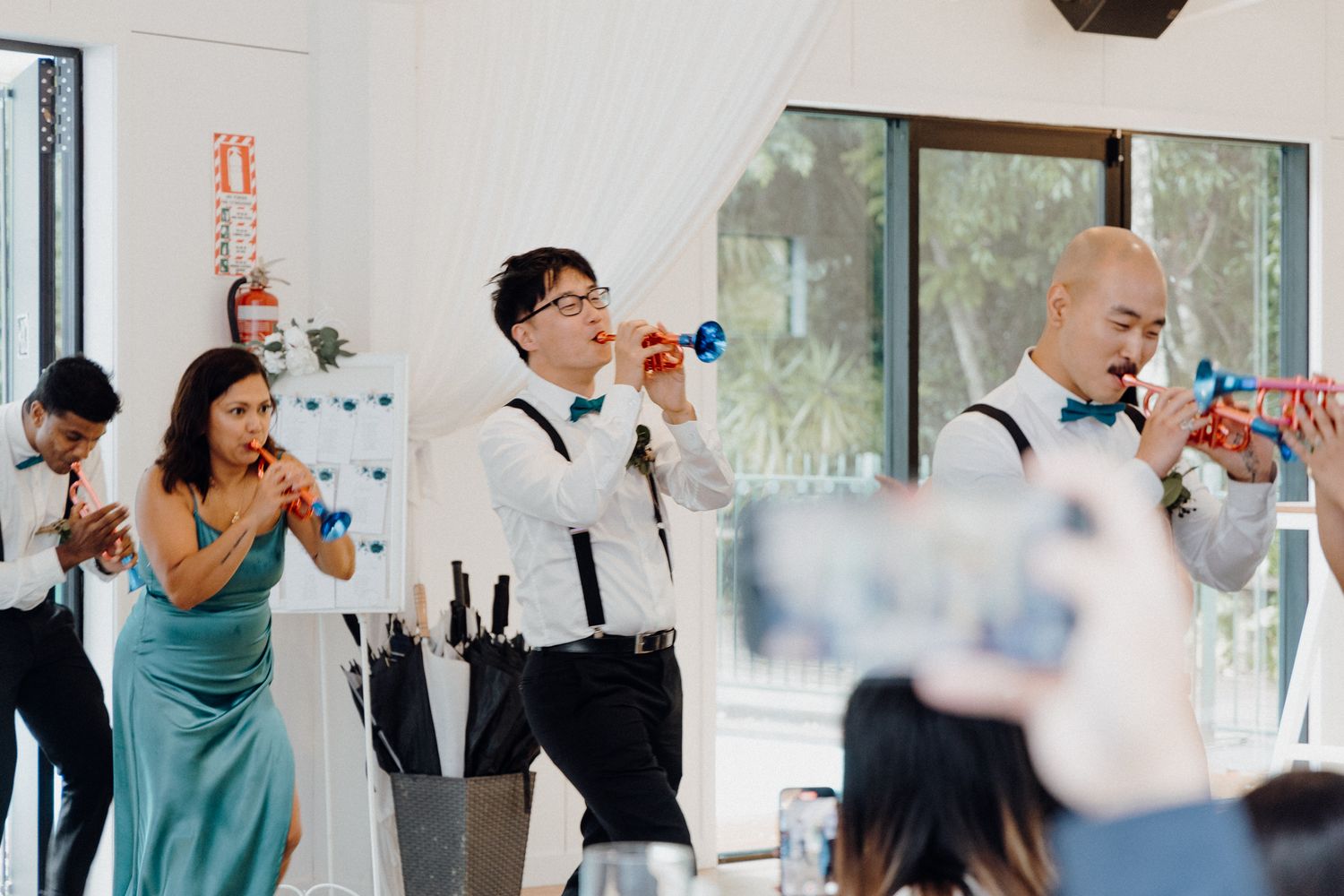 Wedding party attendees in teal dresses and black suspenders cheer captured by Apertured Media Auckland Wedding Photographer.