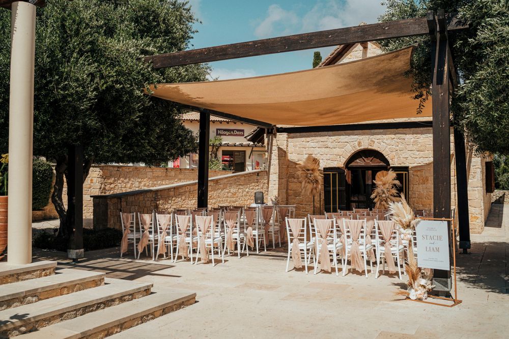An outdoor patio with a tan shade sail and white chairs set up for a wedding ceremony at a rustic stone venue.