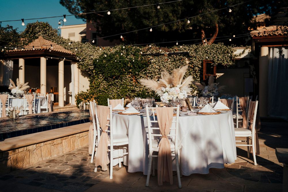 Elegant outdoor wedding reception setup with white tables and chairs arranged on a stone patio under sunlit trees.