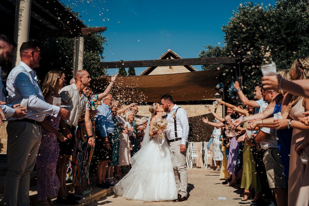 Wedding guests toss confetti over newlyweds exiting ceremony at rustic outdoor venue on sunny summer day.