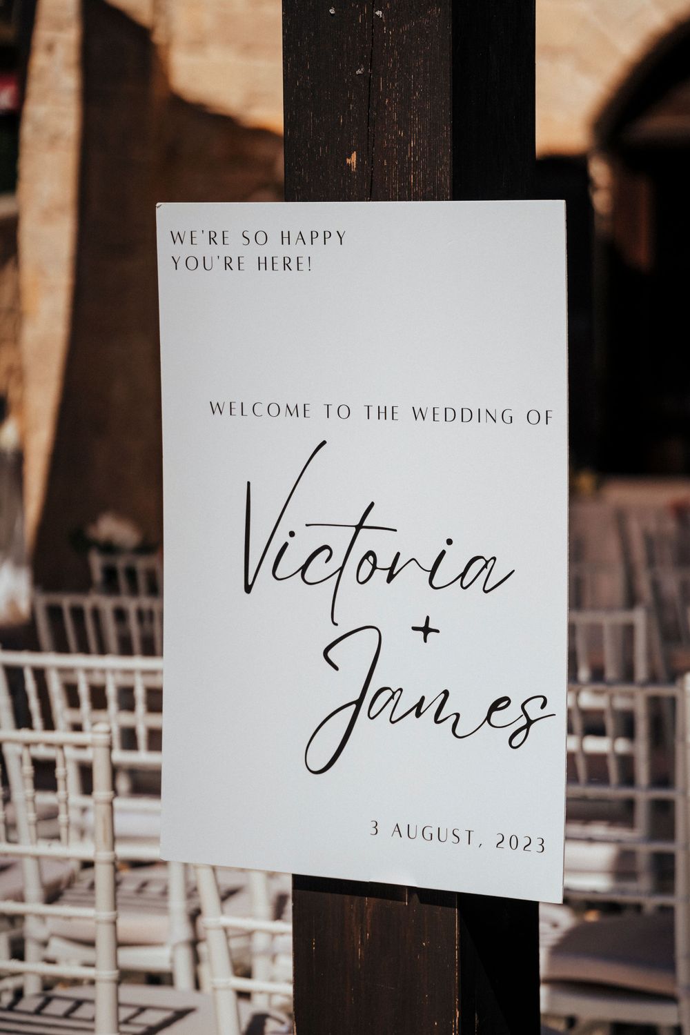 A white wedding welcome sign displays 'Victoria + James' with elegant script text and their August 2023 wedding date.