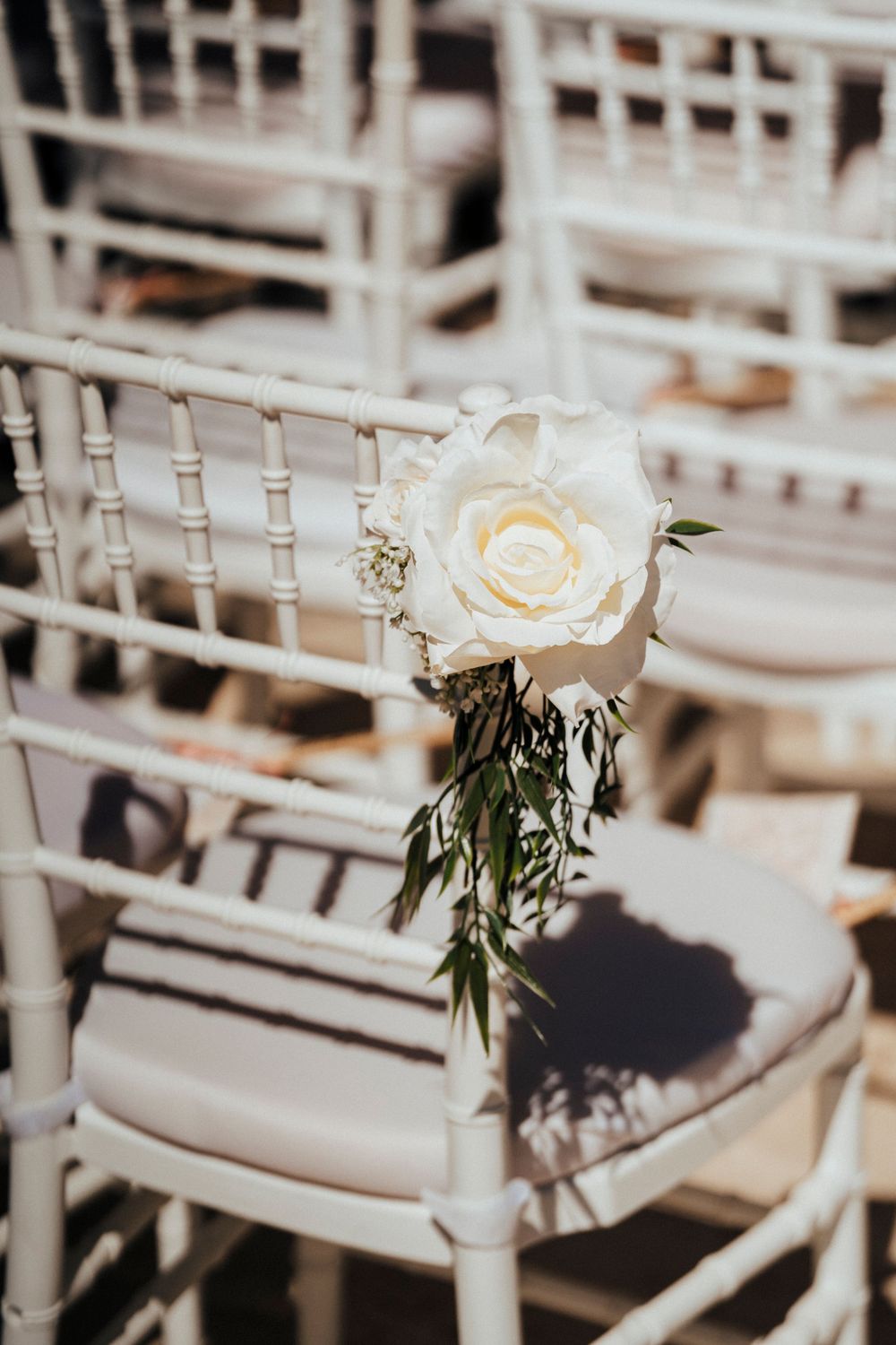A white rose with greenery decorates a wooden chair at a wedding ceremony setup.