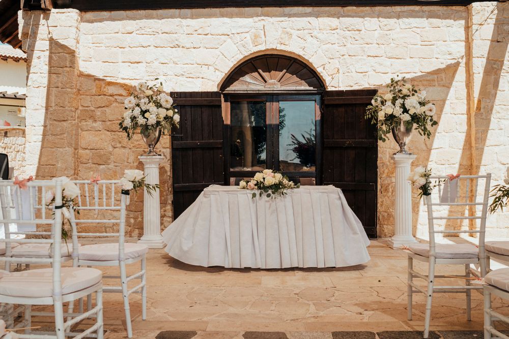 Elegant wedding ceremony setup with white chairs, floral arrangements, and draped table against rustic stone archway entrance.