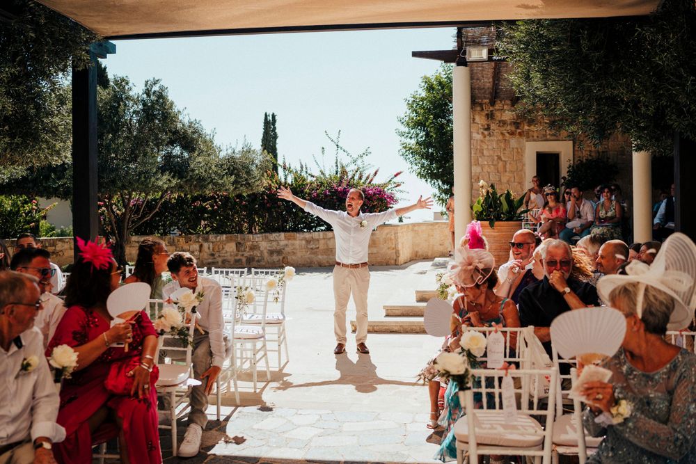 Outdoor wedding ceremony with guests seated in white chairs as someone stands joyfully at the altar during sunset.