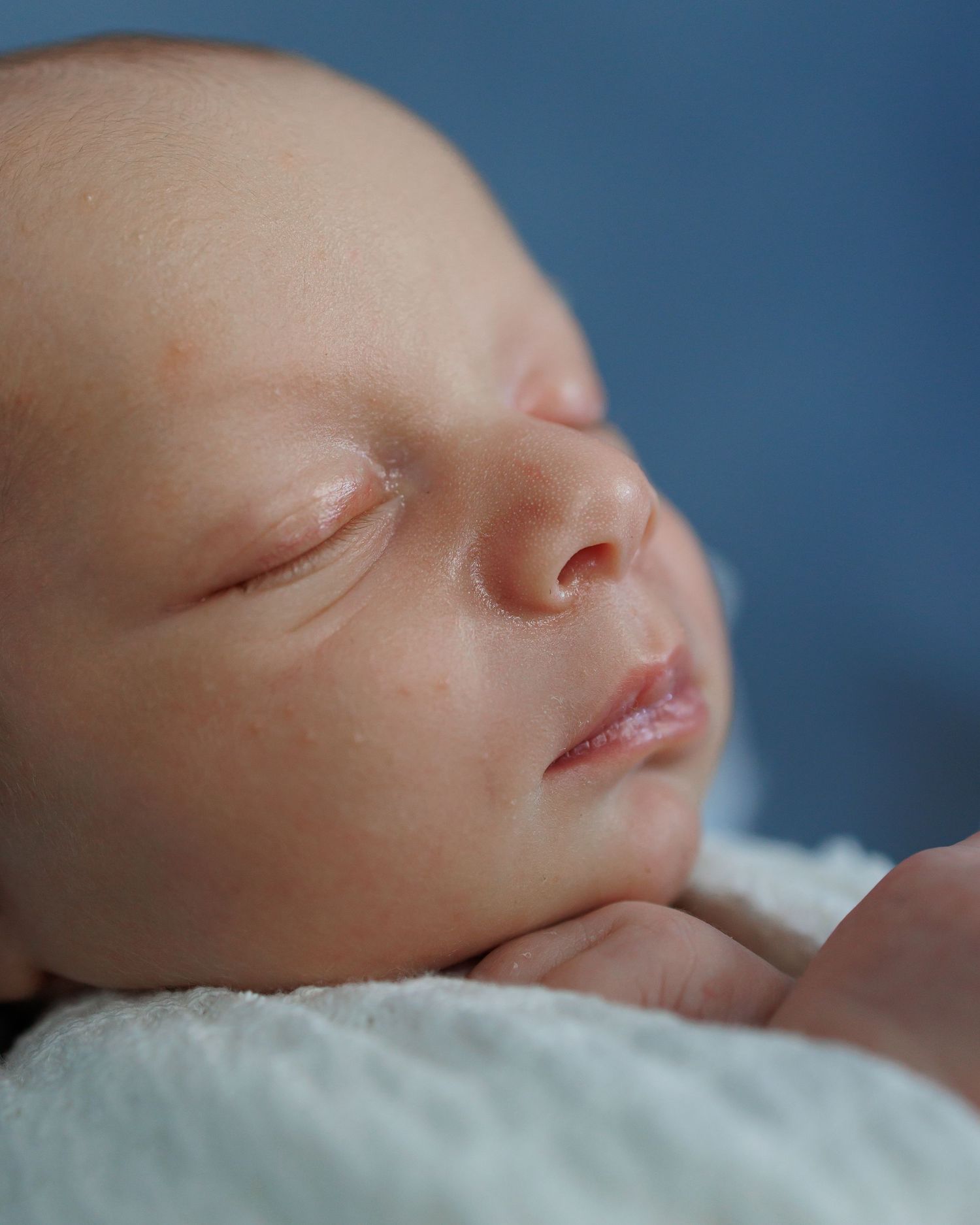 A newborn baby swaddled in white fabric with a matching hat lies peacefully on a brown textured surface.