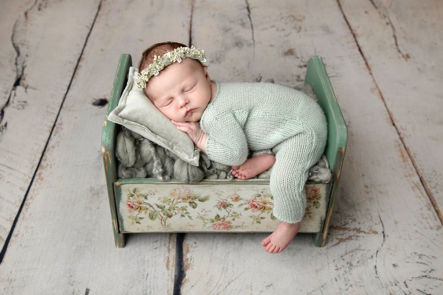 Sleeping newborn in cream outfit nestled in green blanket with lace trim on wooden backdrop.