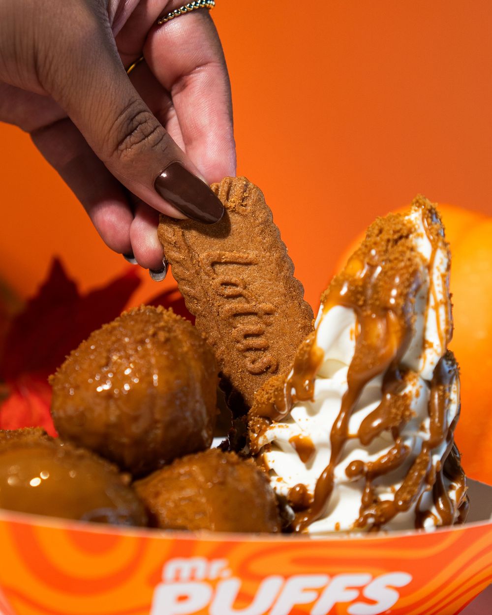 A person dips a chocolate cookie into a swirled ice cream sundae against an orange background.