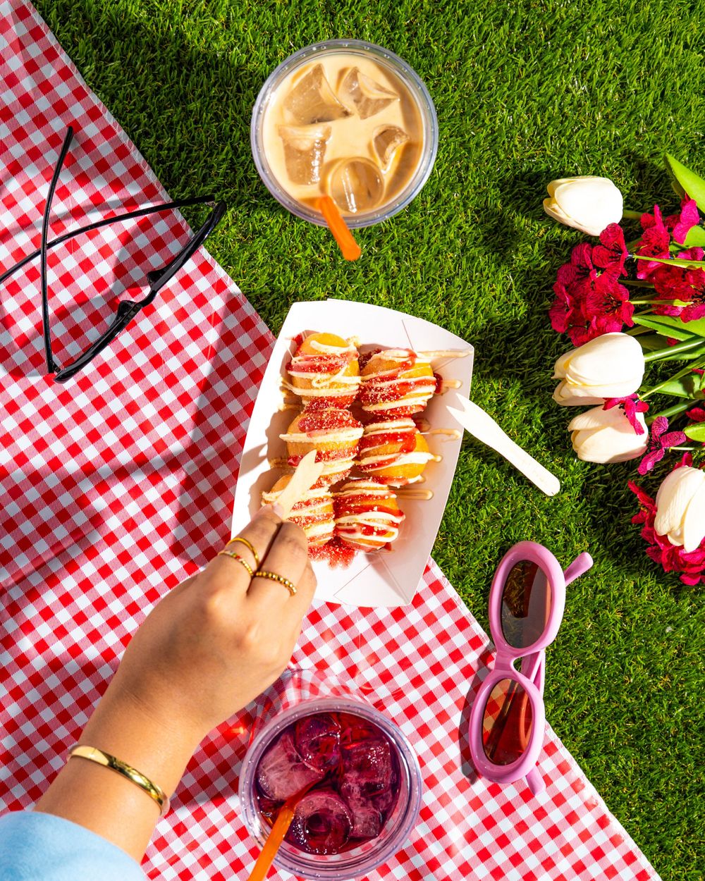 Hands reach for sandwiches on red and white checkered tablecloths with pink flowers and sides on a grassy lawn.