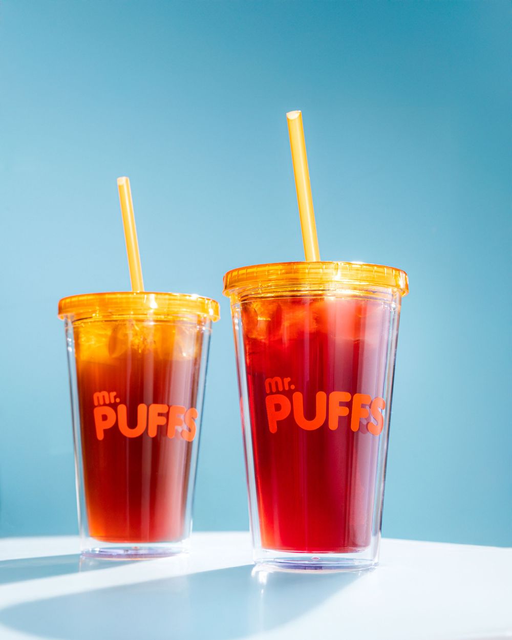 Two clear plastic cups of red iced beverages with orange lids against blue background.Mtlshutter photography