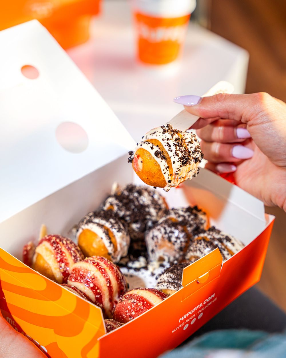 Hand picking up sprinkle-covered pastry from orange branded box filled with assorted treats.Mtlshutter photography