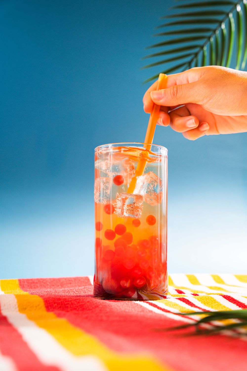 A tropical themed flatlay showing hands pouring orange juice into a glass decorated with palm leaves.