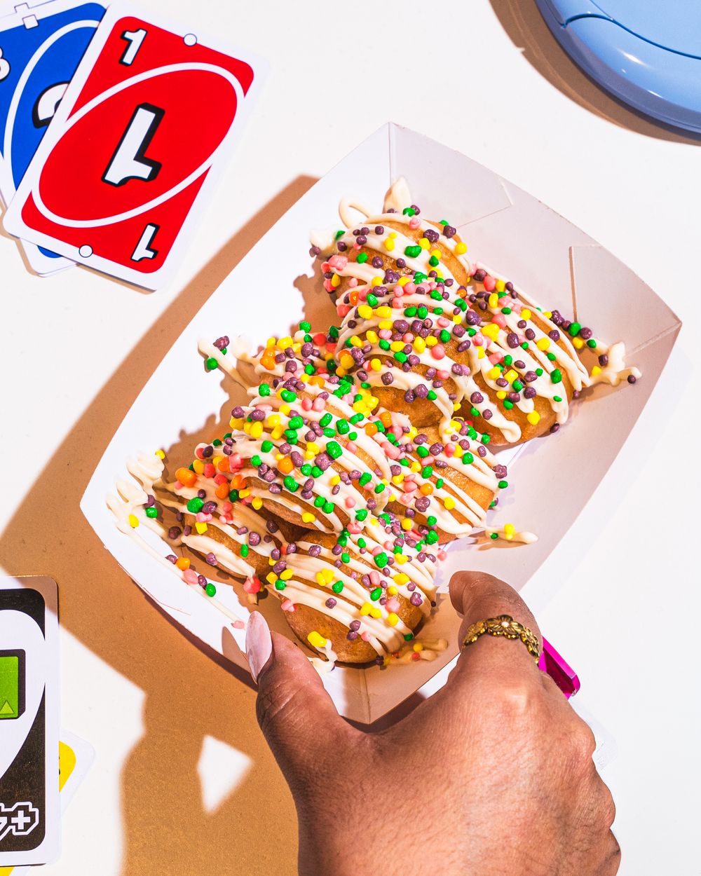 Hand holding a paper tray of churros topped with white icing and colorful sprinkles next to UNO cards.