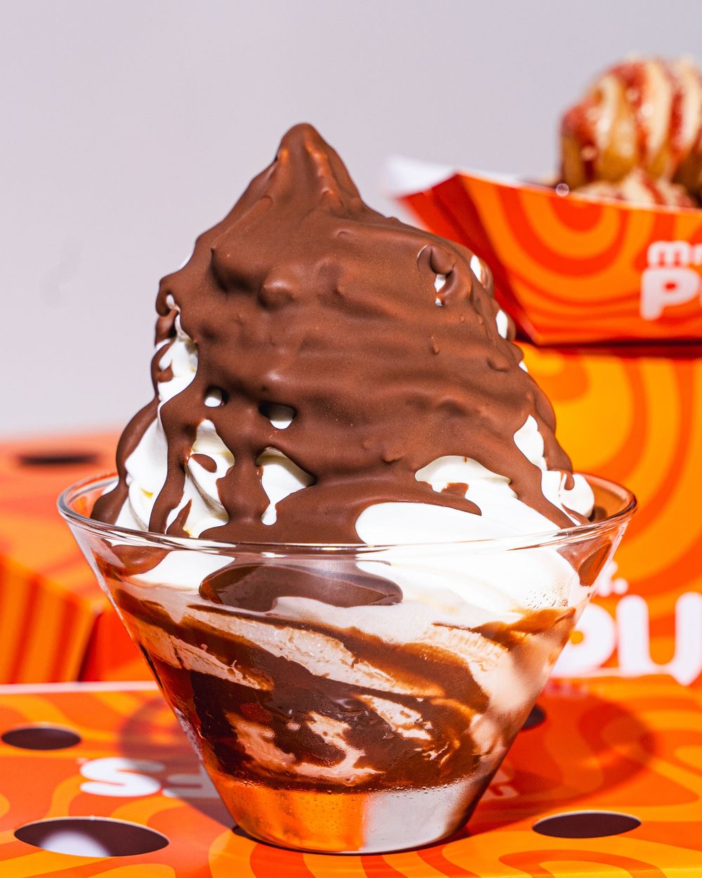 Chocolate soft serve ice cream sundae drizzled with hot fudge sauce in a clear cup on an orange branded tablecloth.