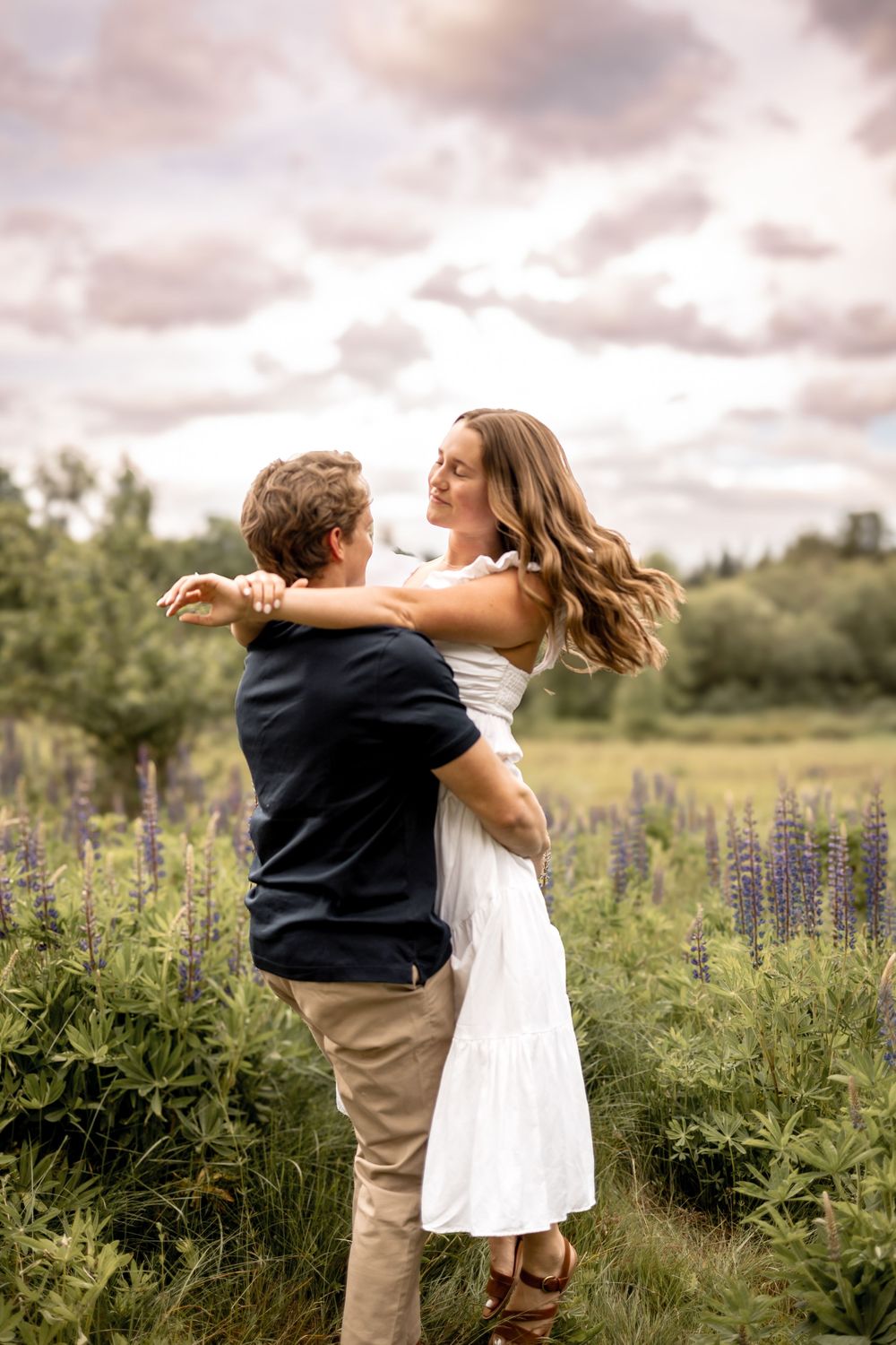 A joyful couple embraces and spins in a field of purple wildflowers at sunset.