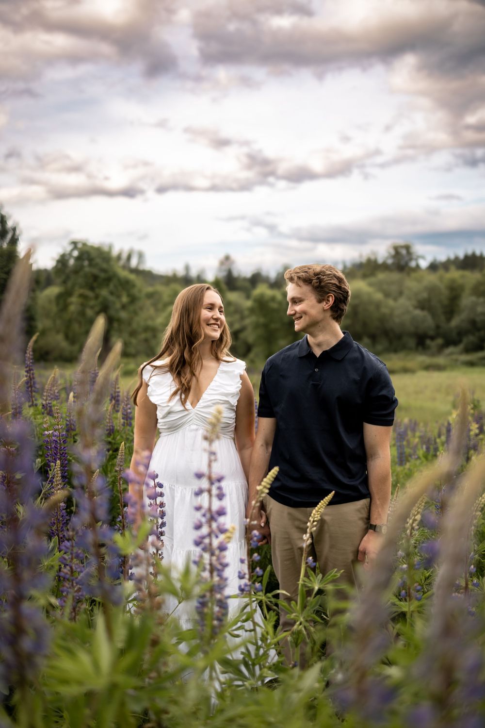 Couple walking through a field of purple lupine flowers under dramatic cloudy skies.