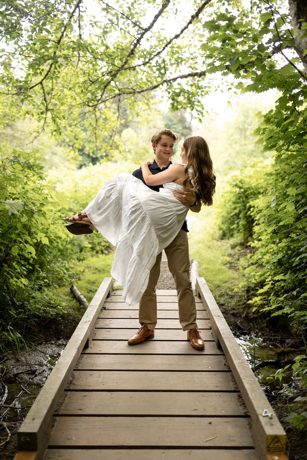 Couple sharing romantic moments on a wooden bridge surrounded by lush green trees.