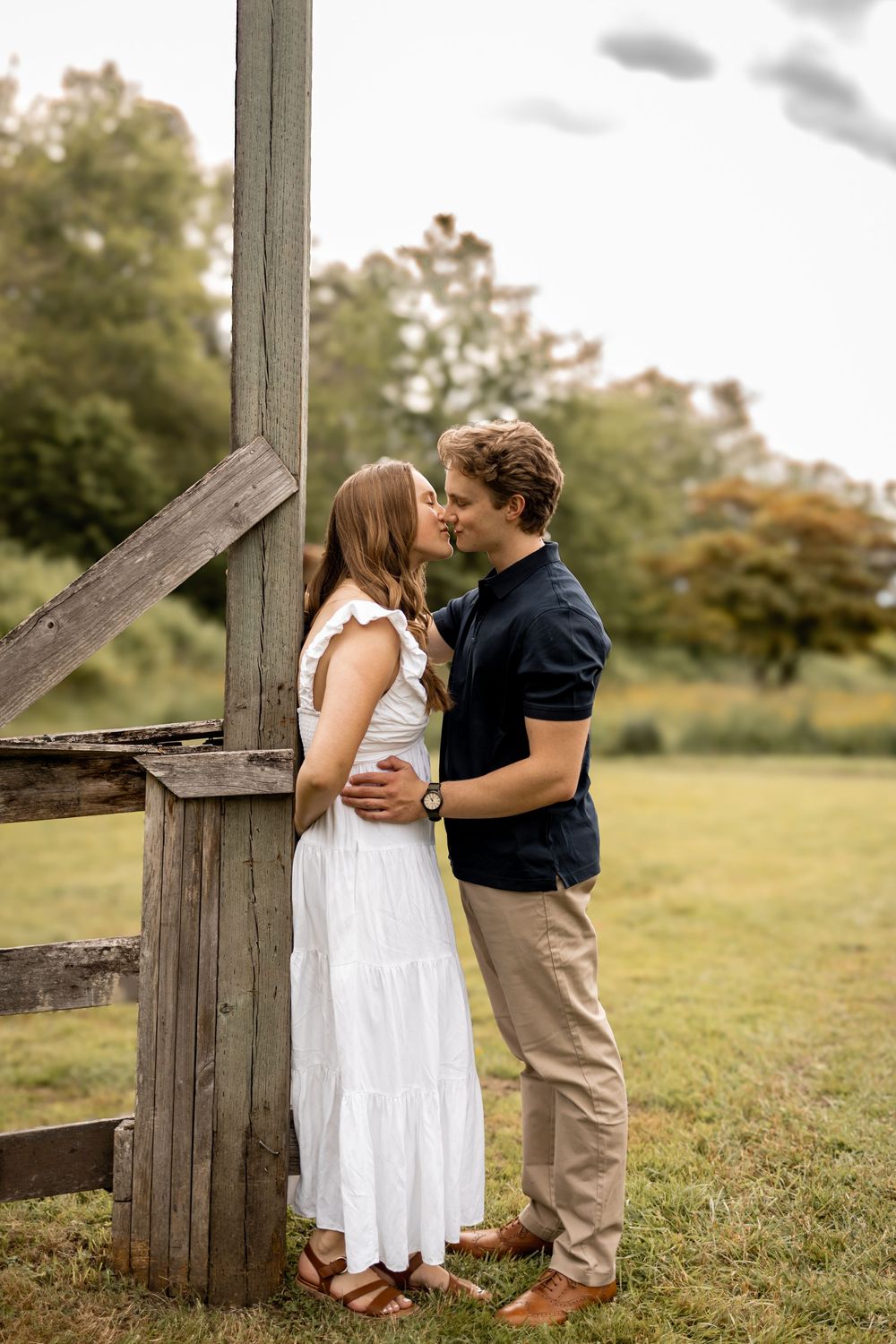 Couple in white dress and casual wear embracing in a grassy field near wooden fence.