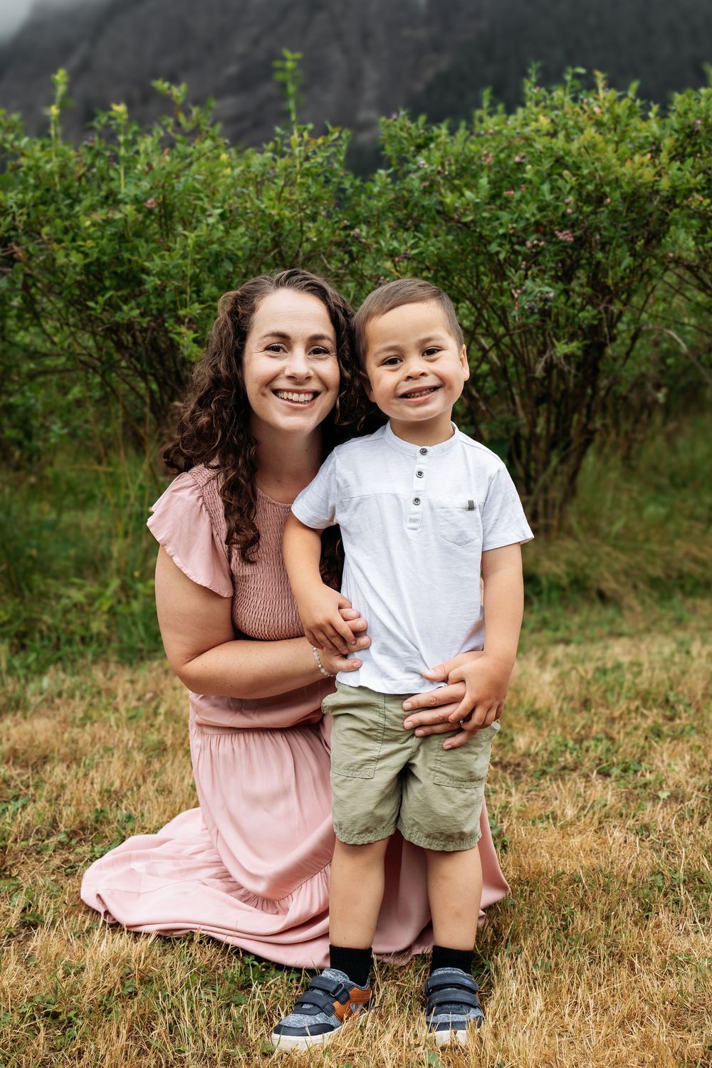 Mother and child sharing a joyful moment outdoors in front of green bushes.