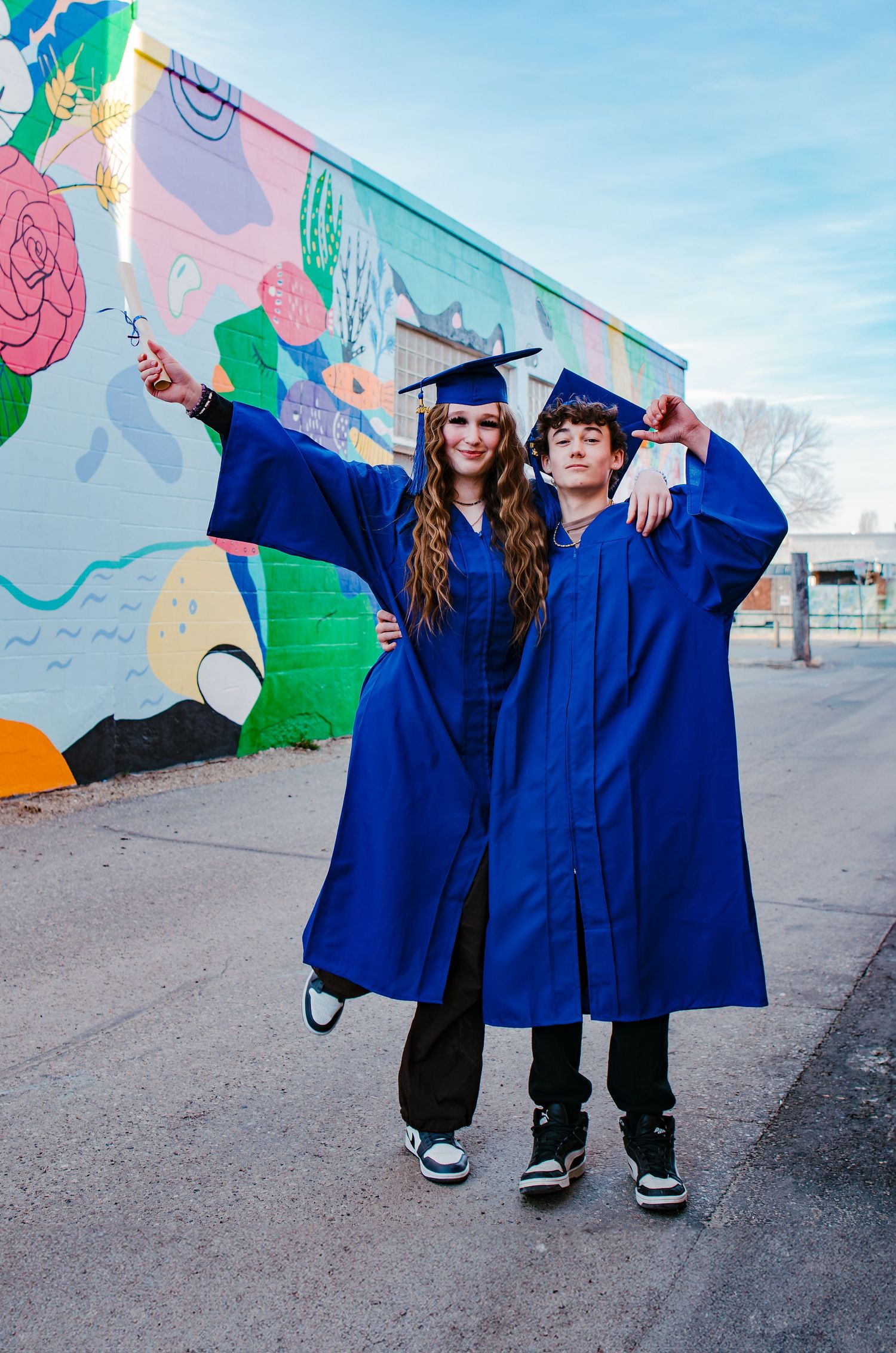 Two graduates in blue caps and gowns pose in front of colorful mural wall.