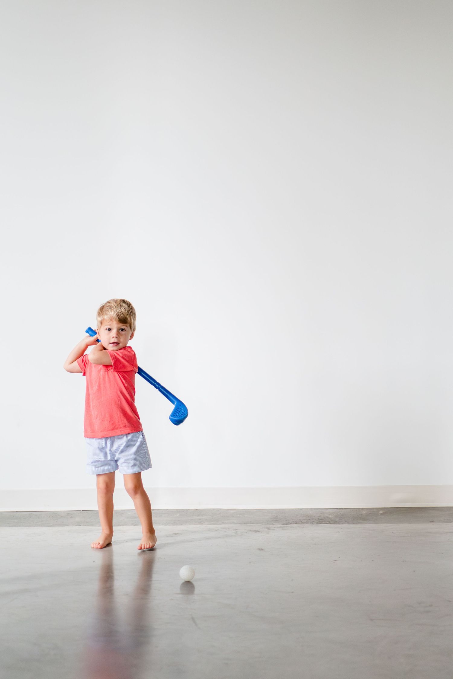 A small child swings a blue golf club while wearing a coral shirt and grey shorts on a bare concrete floor.