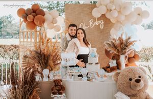 Baby shower decoration setup with balloon arch, pampas grass, and teddy bear in neutral tones.