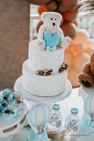 White tiered cake with blue accents and teddy bear topper displayed at a baby shower celebration.