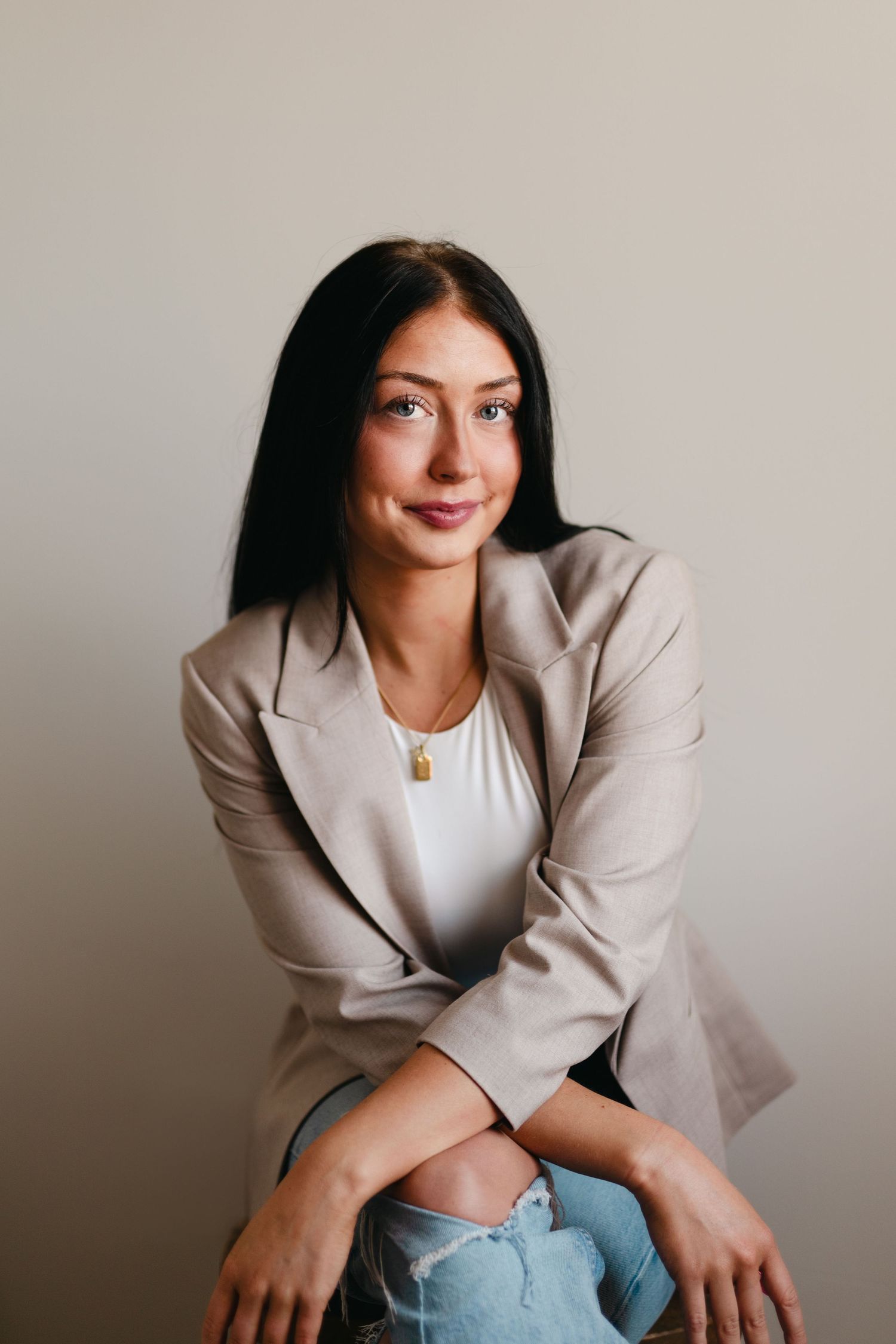 Professional portrait of a person in a beige blazer and white top posing thoughtfully against neutral background.