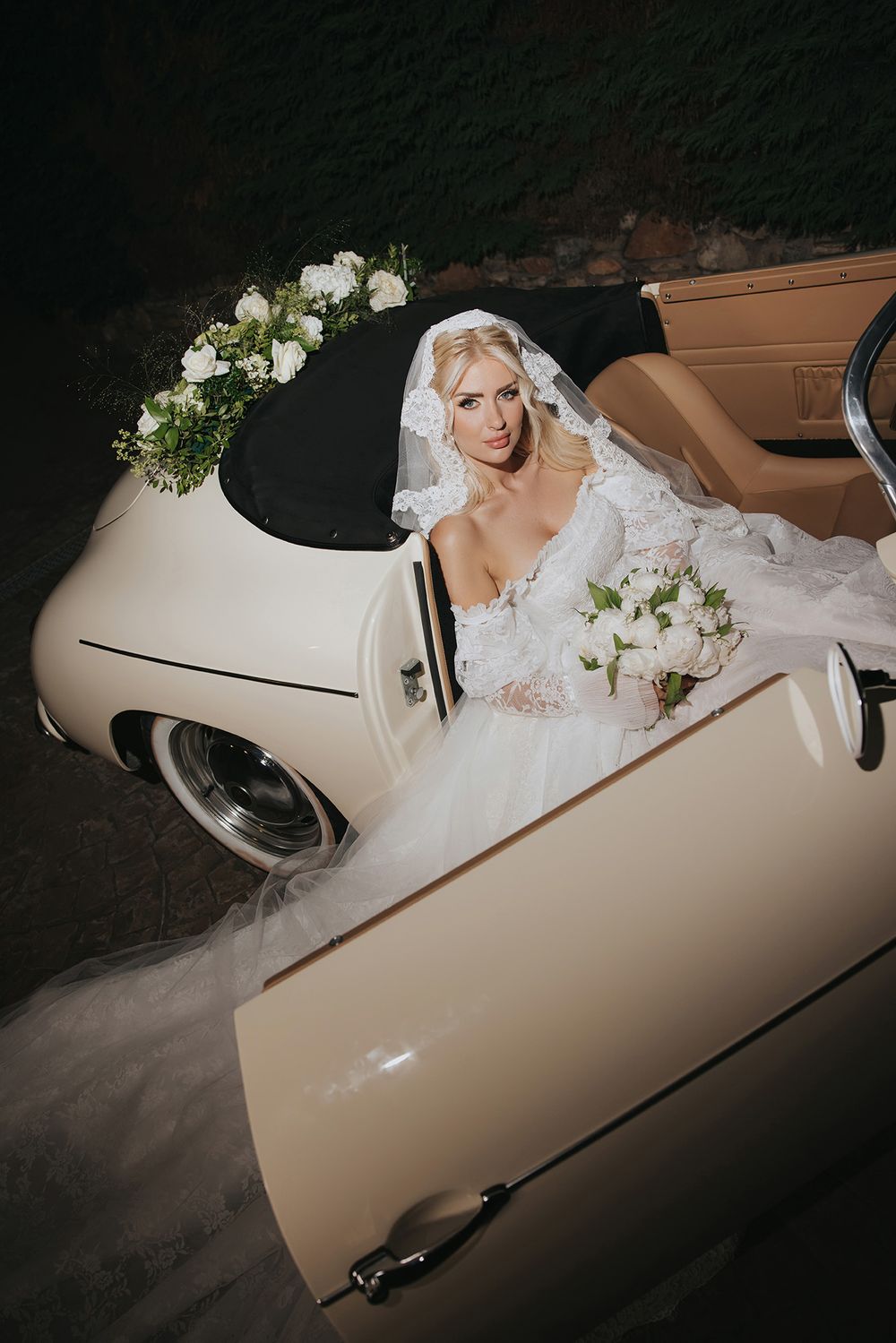 A bride in a white lace gown sits in a vintage cream-colored convertible car decorated with white floral arrangements.