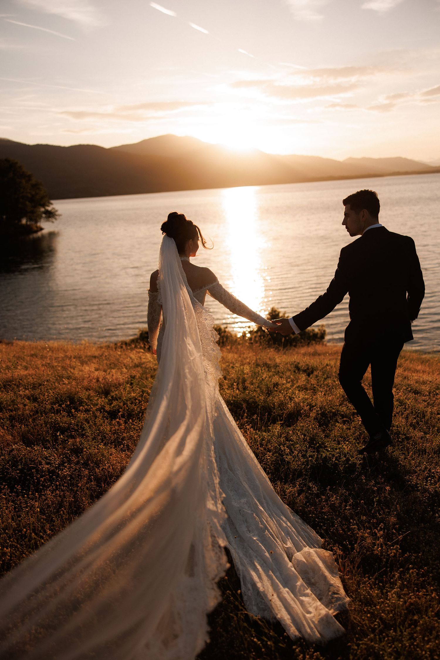 Bride and groom holding hands at sunset by a lake, her long white veil flowing in the breeze.
