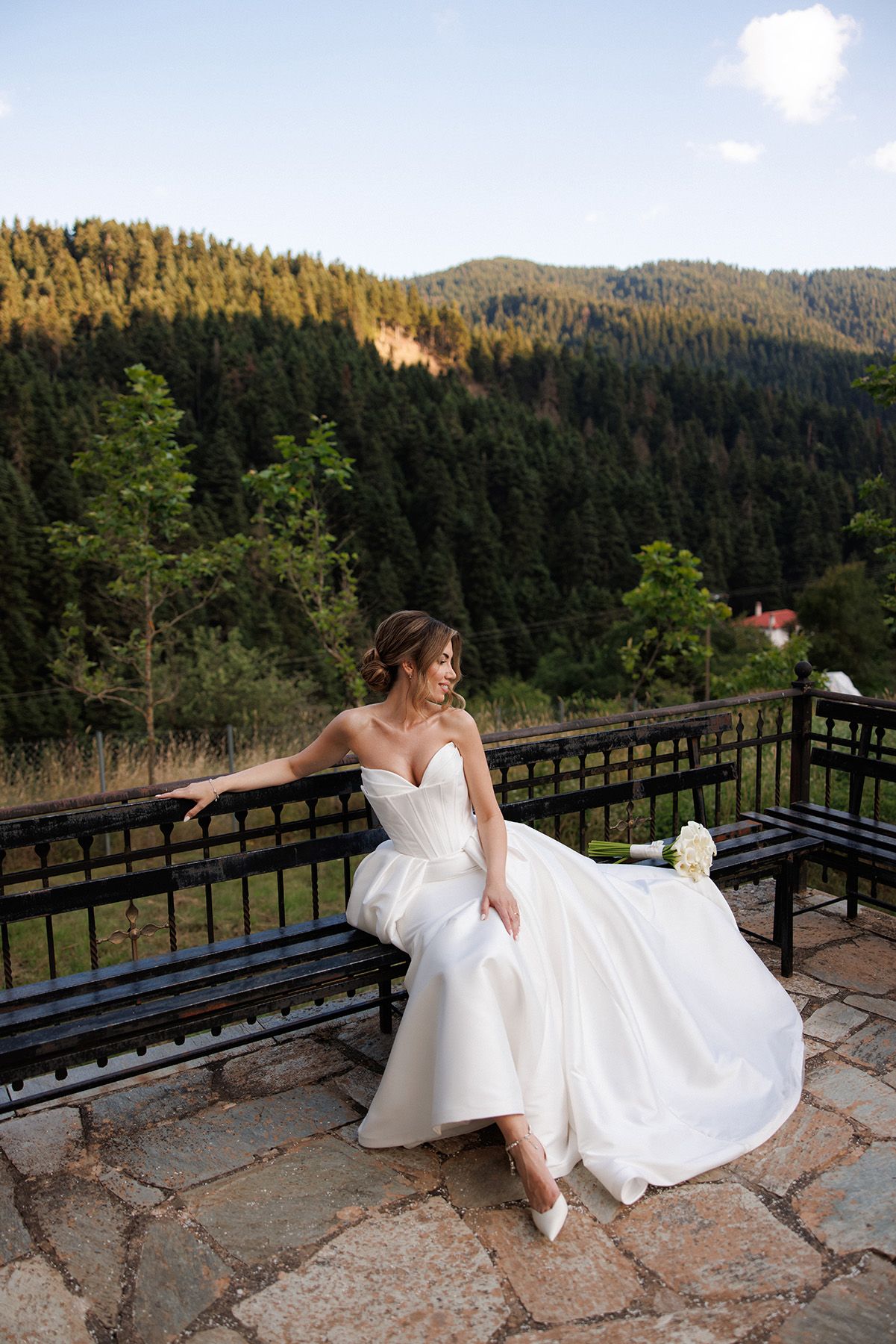 Bride in white strapless gown sits on bench at mountain terrace with lush forest backdrop.