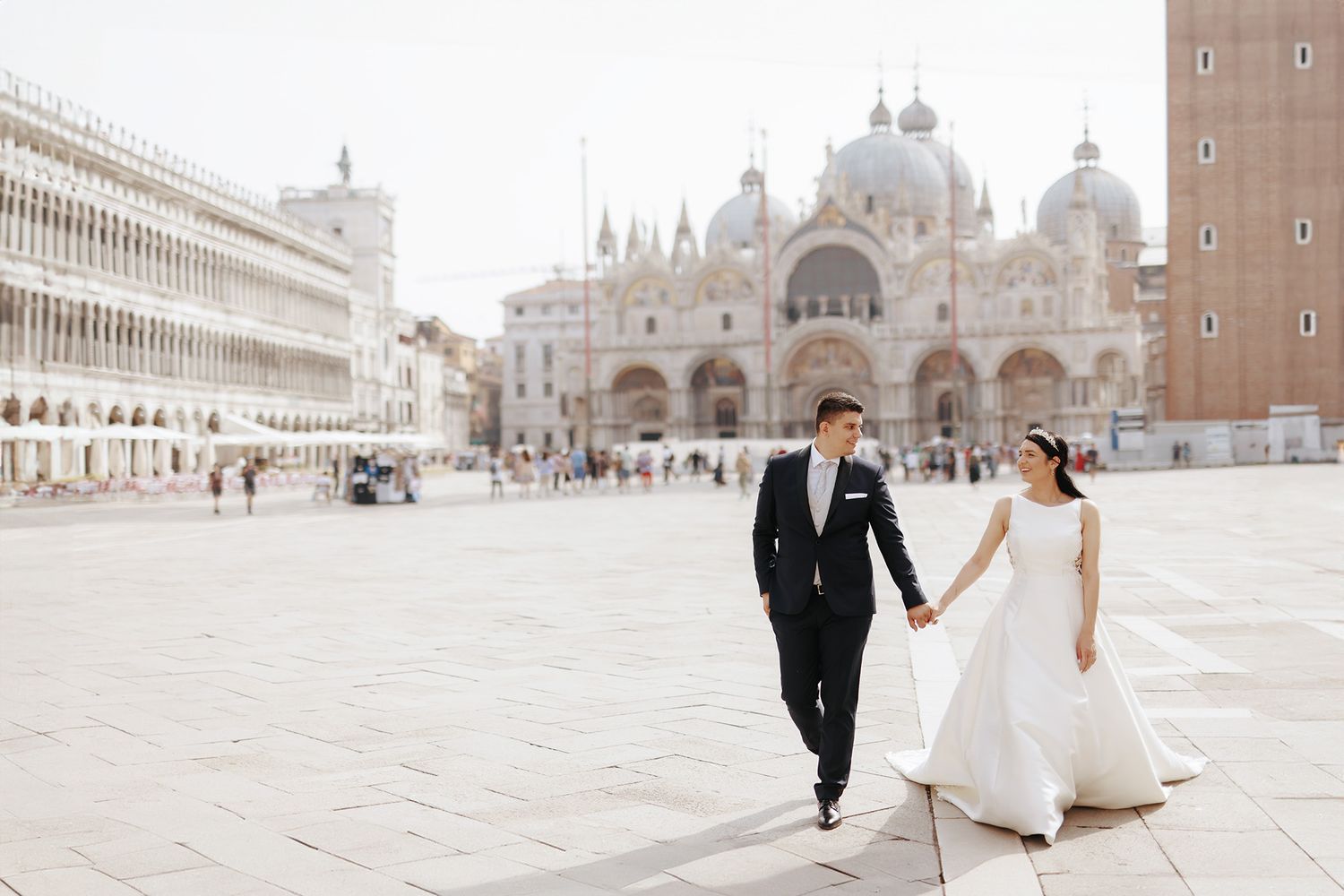 A couple in wedding attire walks across St. Mark's Square in Venice, Italy, with historic architecture in the background.