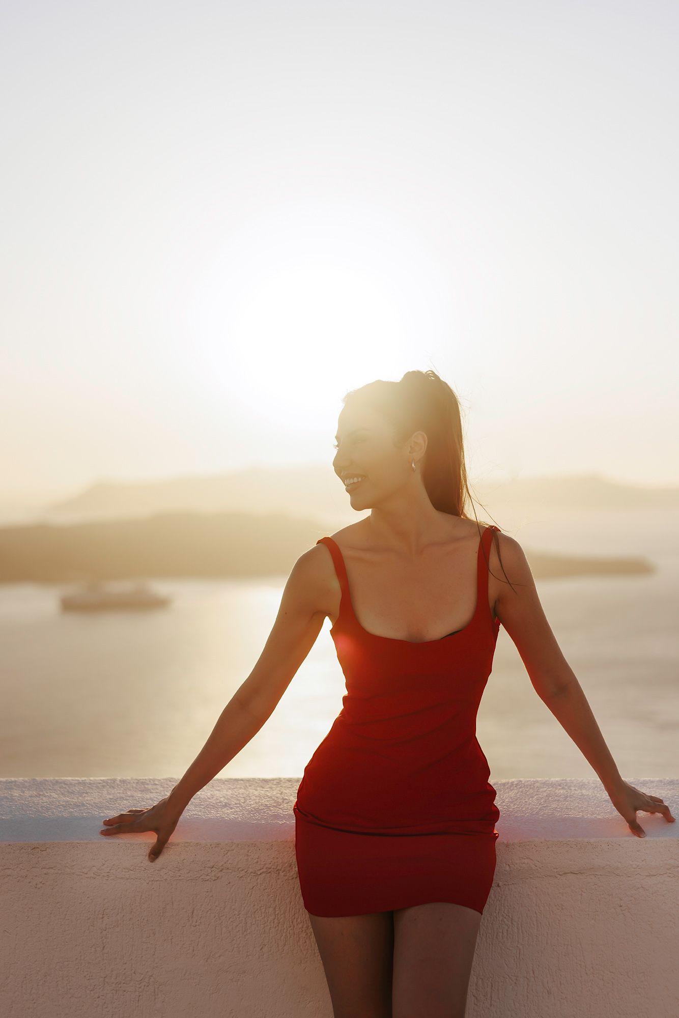 Silhouette of person in red dress admiring scenic ocean view at sunset with boats in distance.