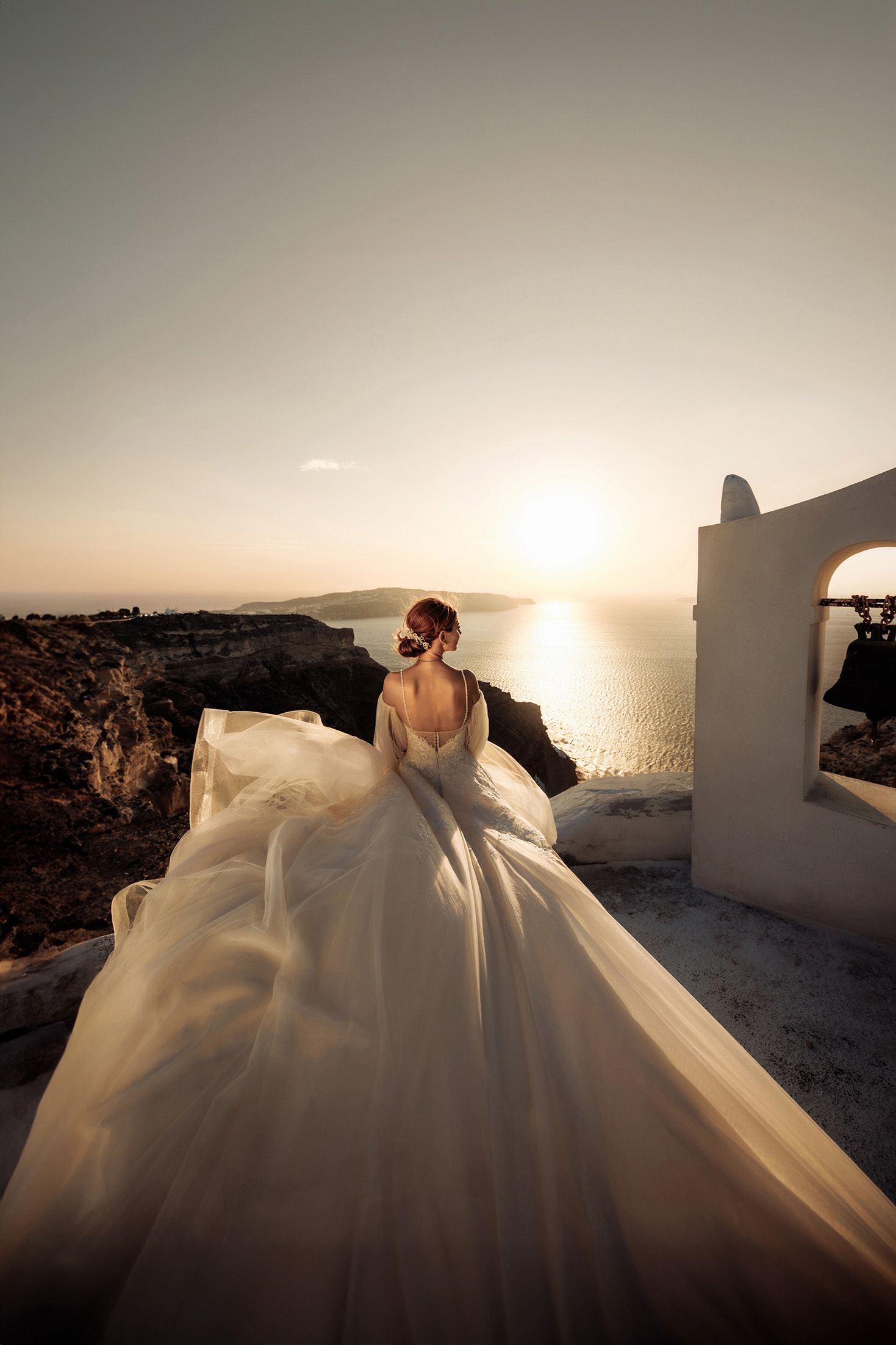 A flowing white wedding dress billows in the wind against a stunning sunset over the Mediterranean Sea in Santorini.