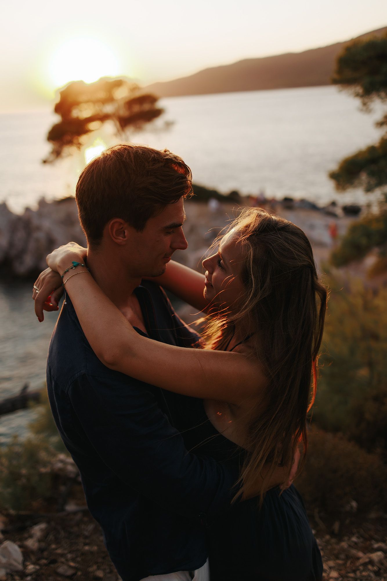 A romantic couple embraces against a golden sunset backdrop among rocks and trees.
