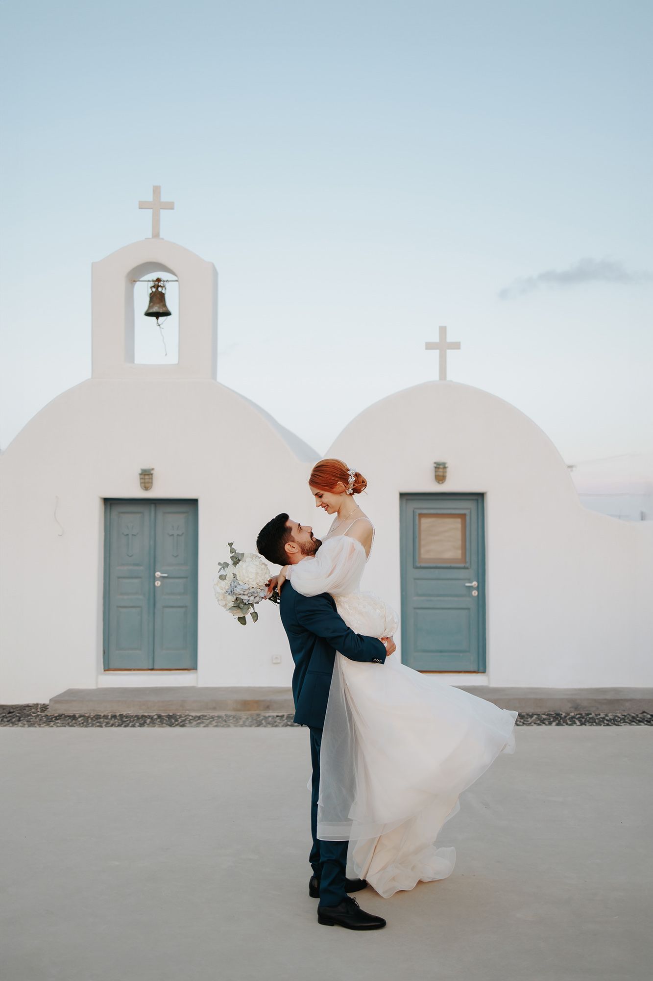 A wedding couple embraces in front of a traditional white Greek church with blue doors and bell tower in Santorini at dusk.