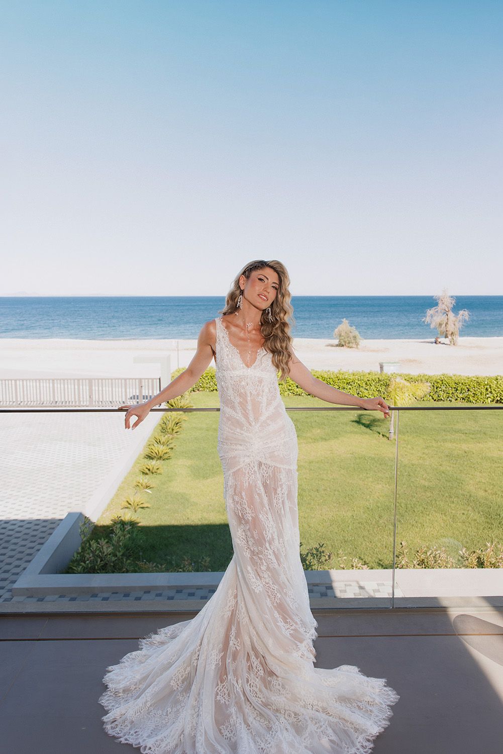 Bride in lace mermaid wedding gown poses on oceanview balcony with beach and blue sky backdrop.