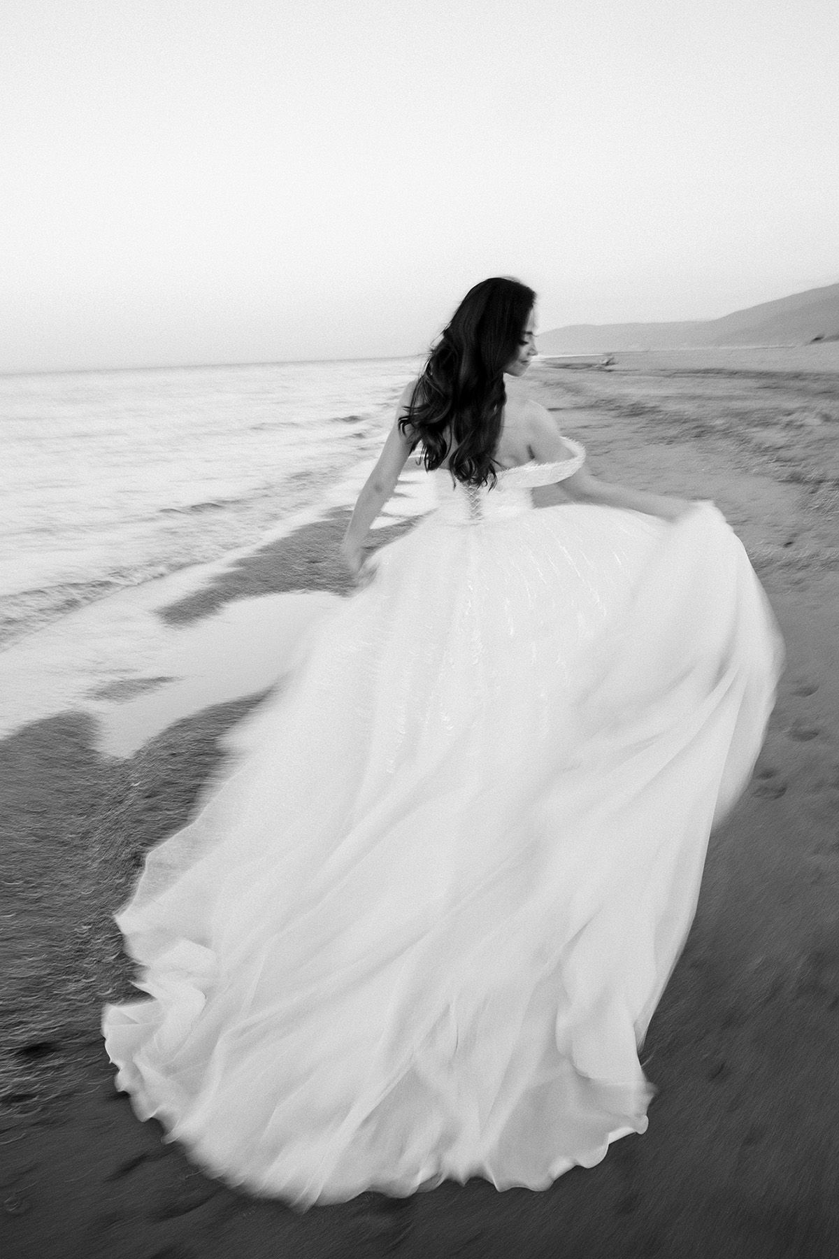 A flowing white wedding dress billows dramatically against a dark beach shoreline in a black and white photo series.