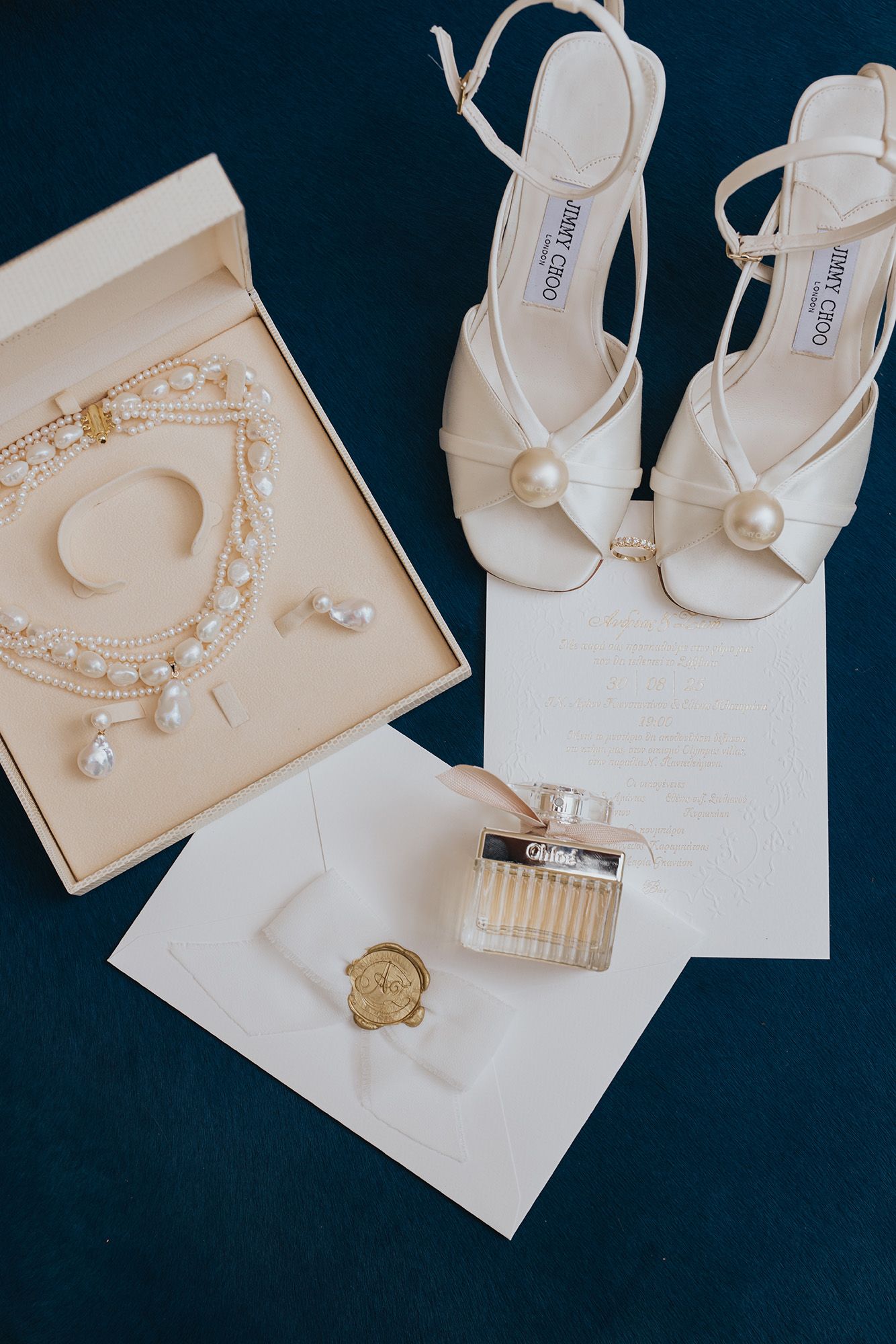 White wedding accessories including pearl jewelry, delicate sandals, and elegant stationery arranged on dark background.