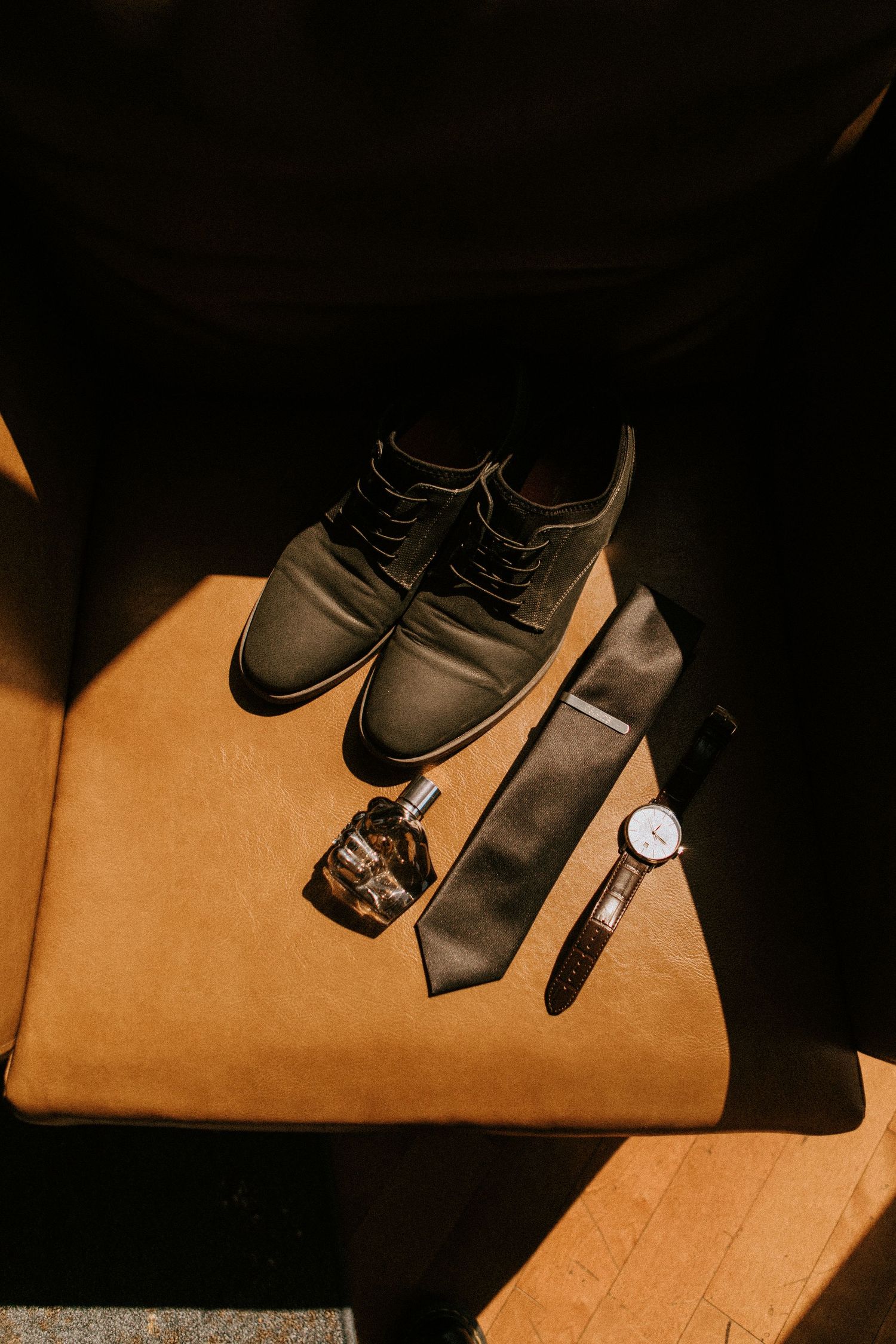 Wedding accessories including black dress shoes, tie, watch and cologne on tan surface.