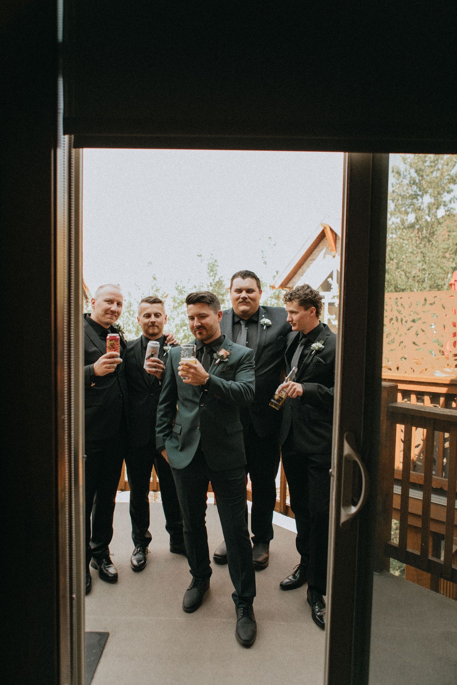 Groomsmen in dark suits gathered on a porch sharing drinks and laughter during wedding celebrations.