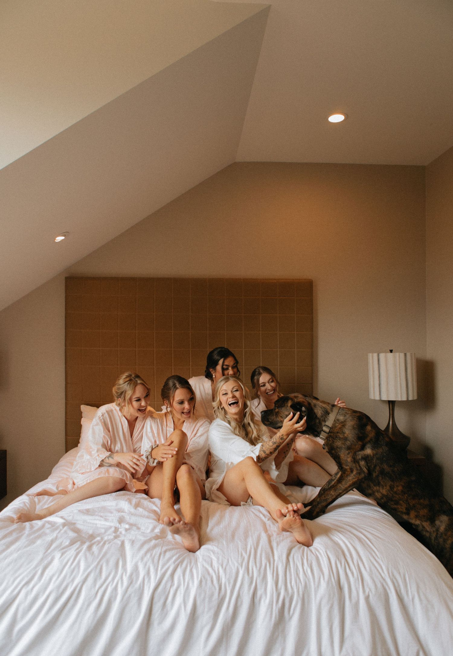 Wedding party relaxes on white bed with dog during pre-ceremony preparations.