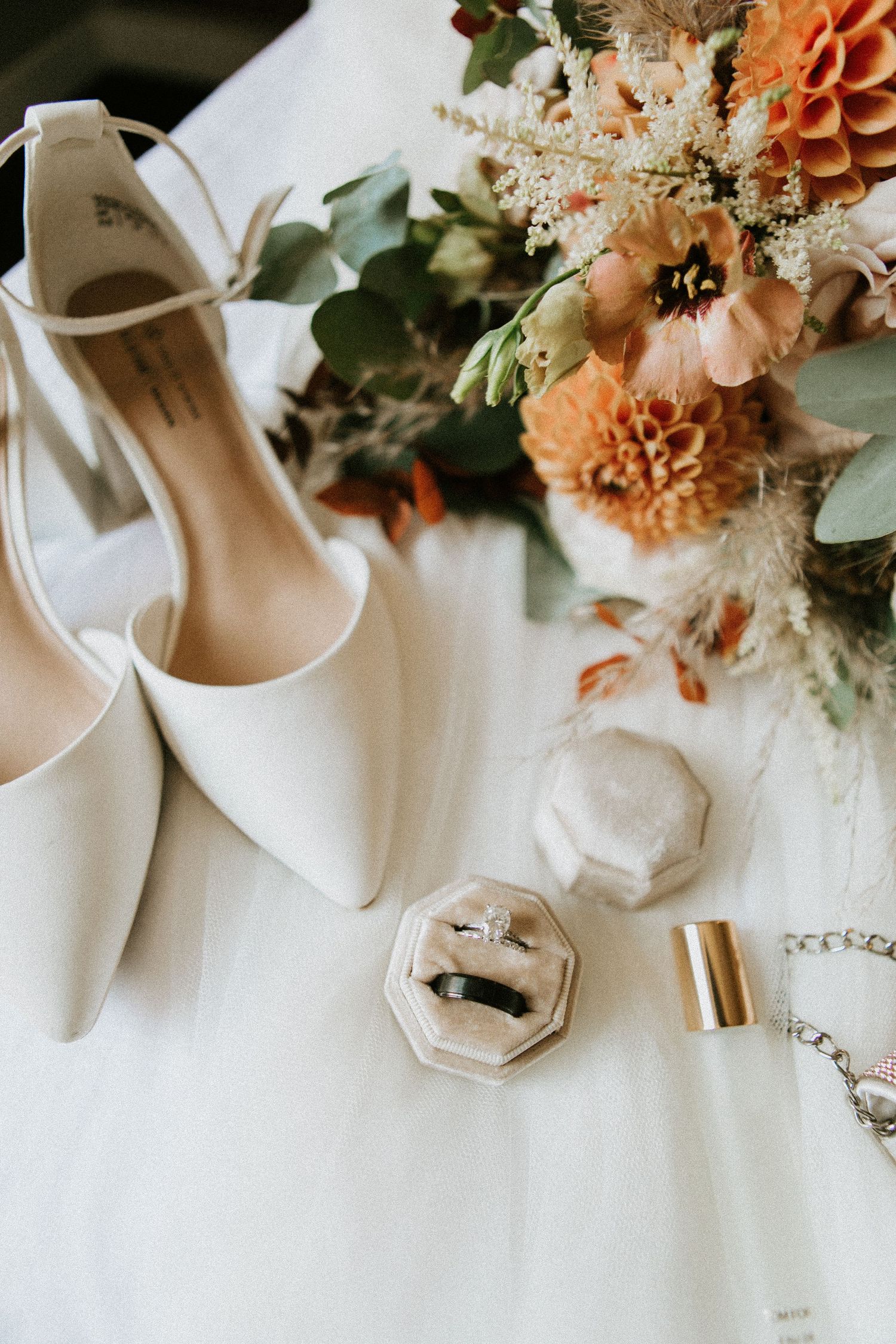 White bridal shoes and wedding jewelry alongside peach and coral floral arrangements on white fabric.