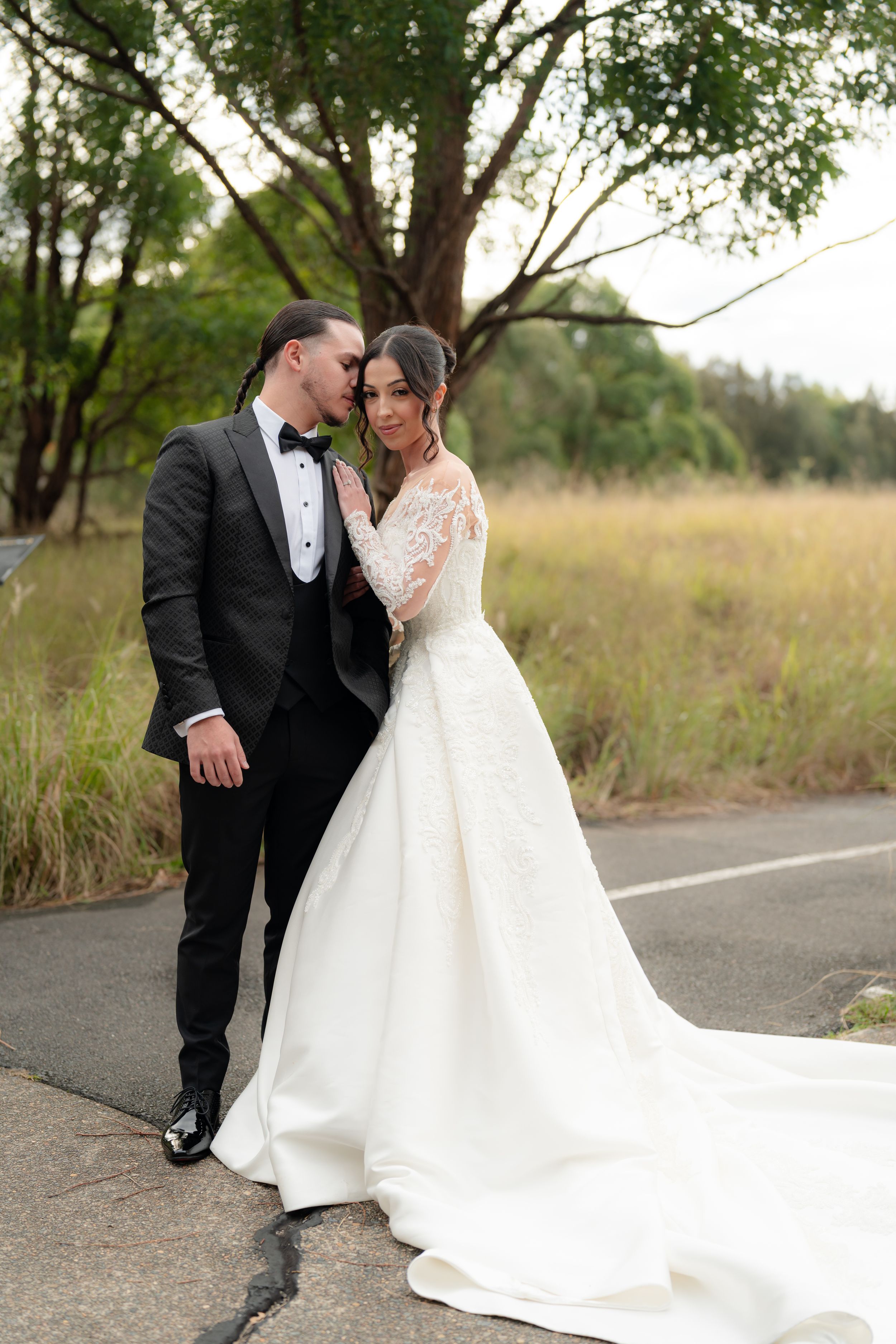 Bride in long-sleeve lace wedding gown and groom in black tuxedo share an intimate moment on a scenic road.