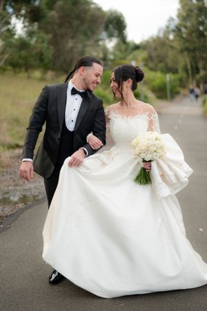 Smiling bride and groom walk together on a path, groom helping carry the bride's elegant white ball gown train.