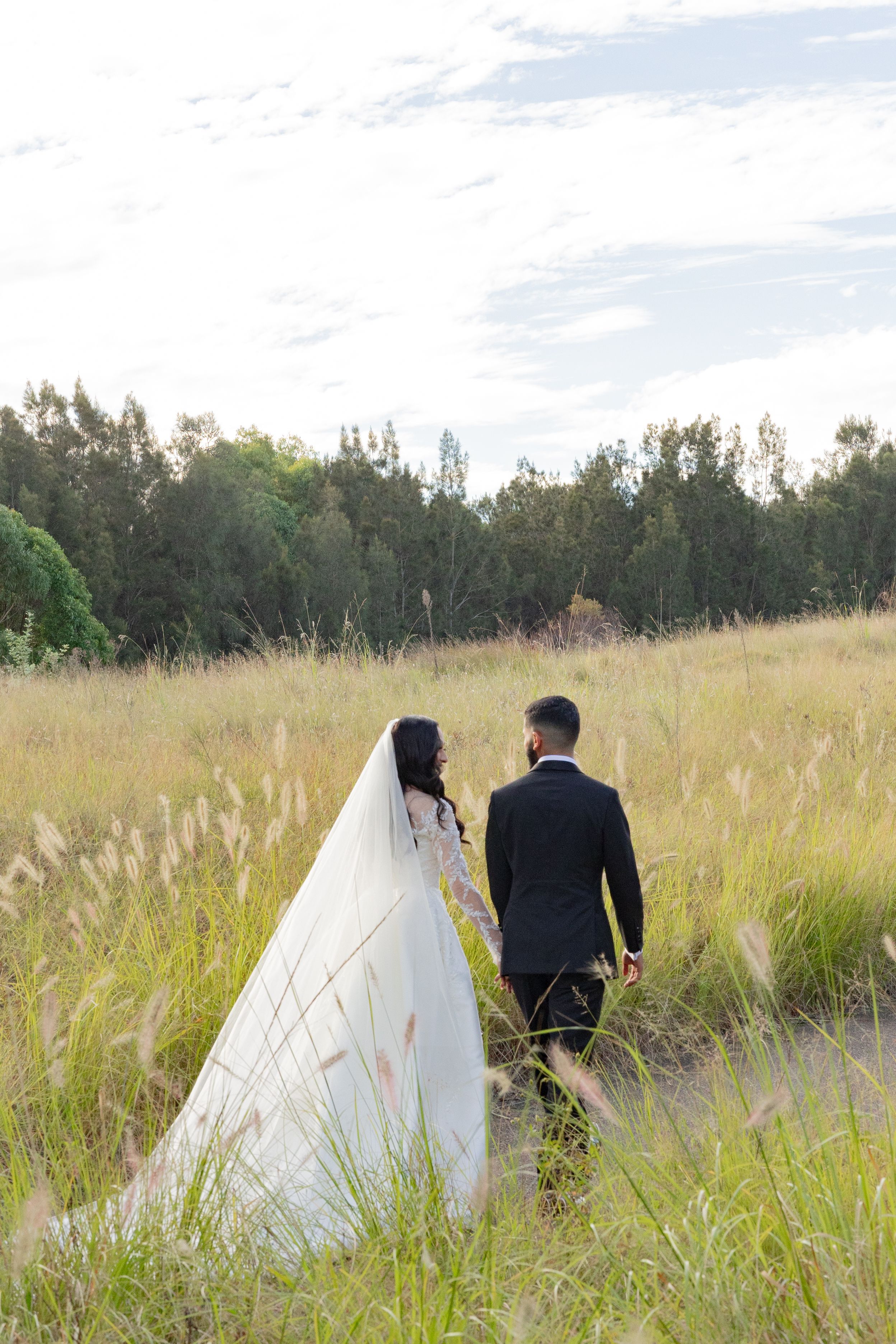 Bride and groom walk hand in hand through golden meadow grass with a lush forest backdrop on their wedding day.