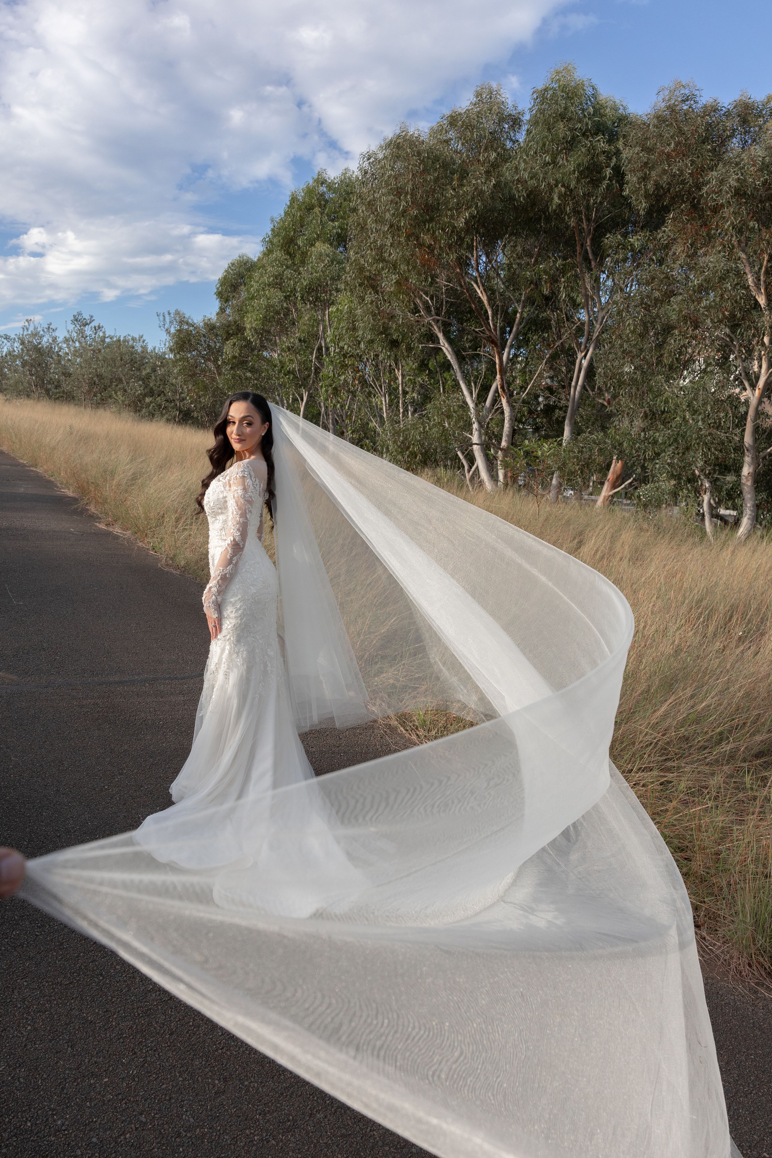 Bride in long-sleeve lace wedding gown poses on a road with her flowing veil billowing dramatically in the wind.
