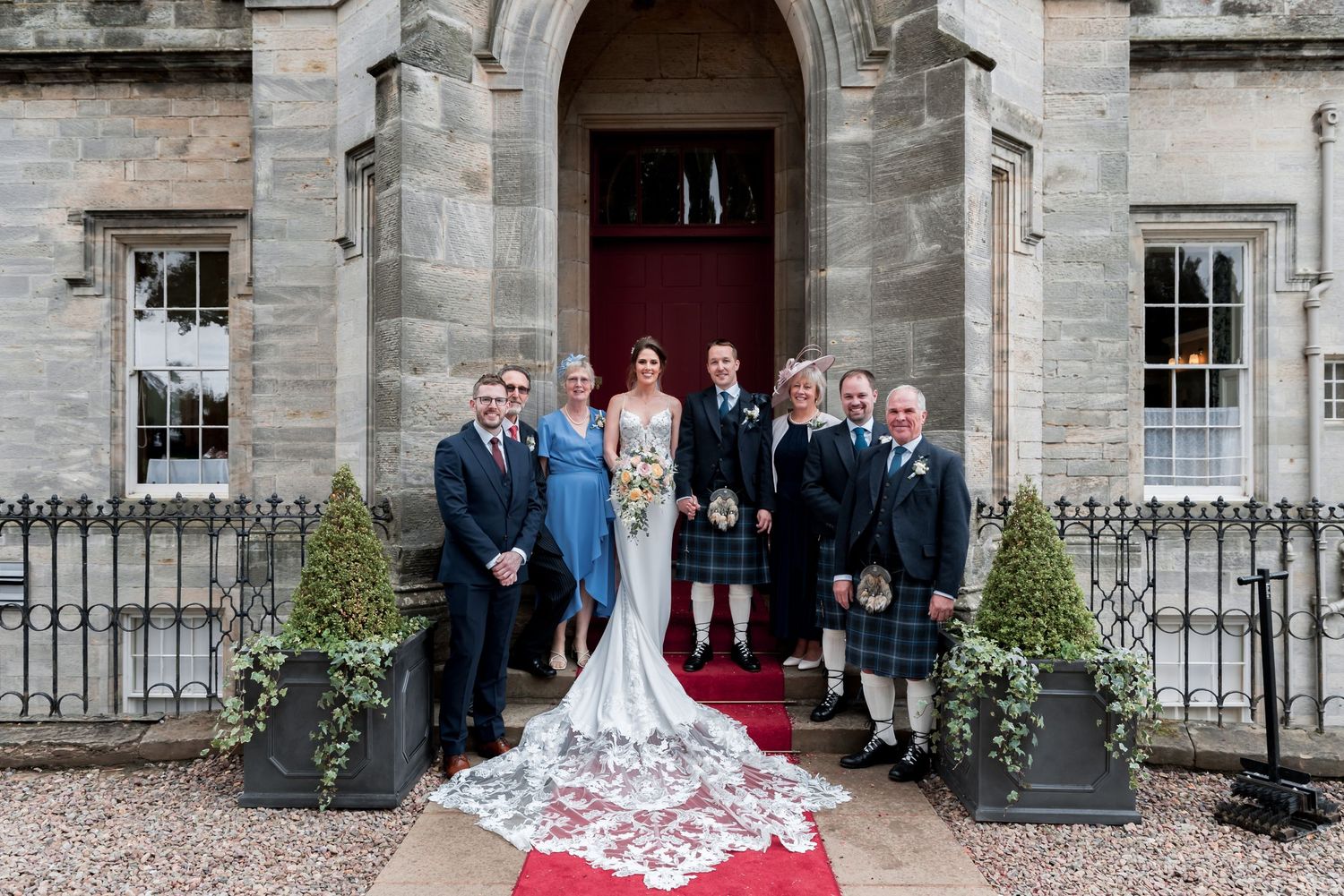 Wedding party gathered on red carpet outside grand stone building entrance with ornate carved doorway.