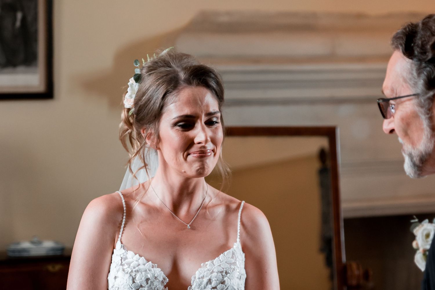 Bride in lace wedding dress and upswept hair stands chatting during indoor celebration.
