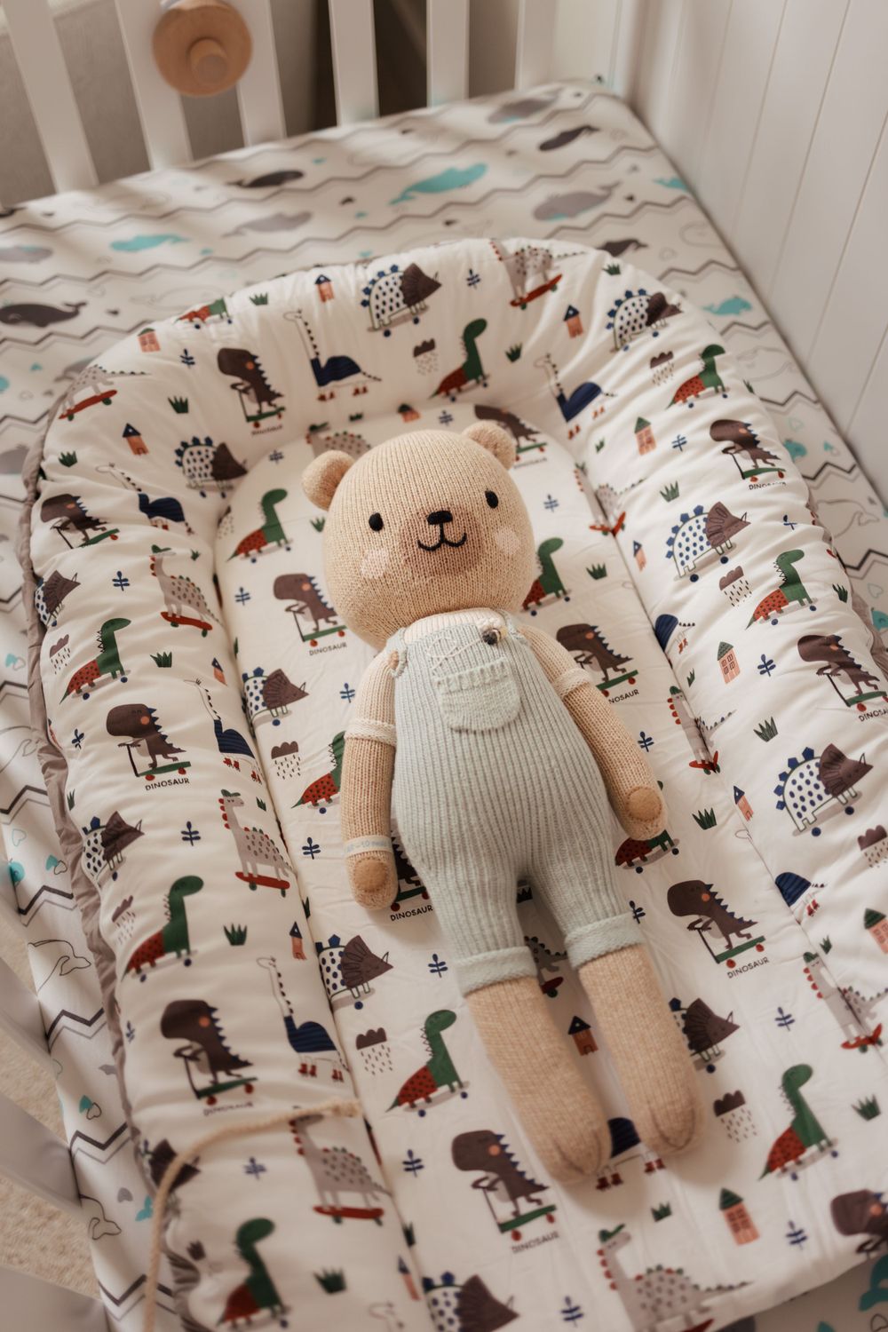 A soft teddy bear toy wearing gray overalls lies on a patterned animal print crib sheet.
